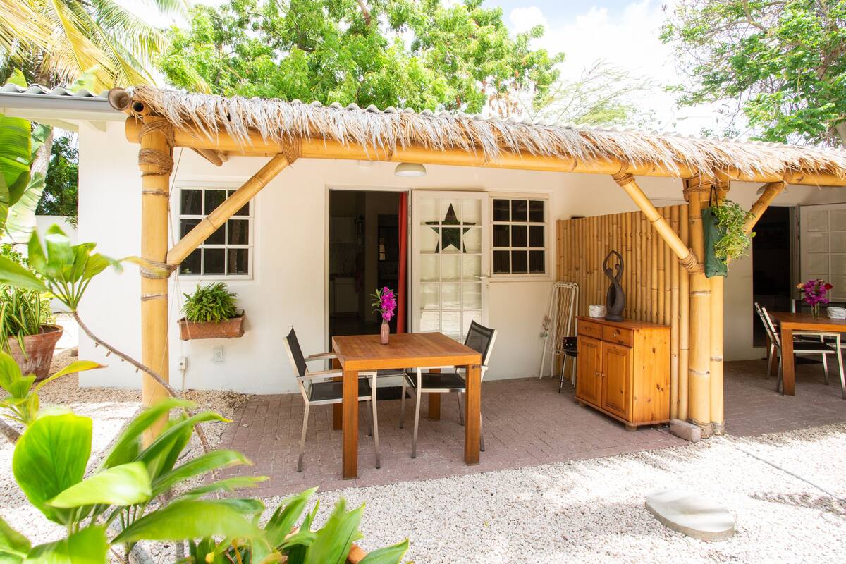 A rustic outdoor dining area with a thatched roof, wooden table, and chairs.