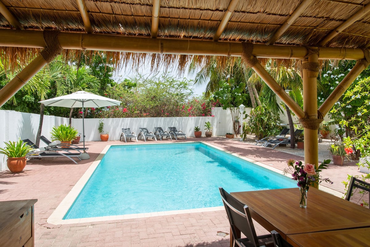 A swimming pool with lounge chairs and umbrellas, surrounded by potted plants and a white fence.