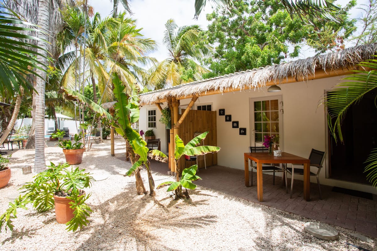 A thatched-roof structure with a table and chairs, surrounded by potted plants and palm trees.