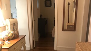 Un couloir doté d’un plancher en bois, d’un tapis à motifs, d’une chaise en osier et d’une commode surmontée d’une lampe décorative.