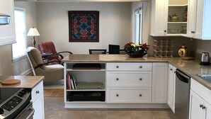 A modern kitchen with white cabinetry, a built-in oven, and a dishwasher. There is a dining area with a table and chairs, and a window providing natural light.