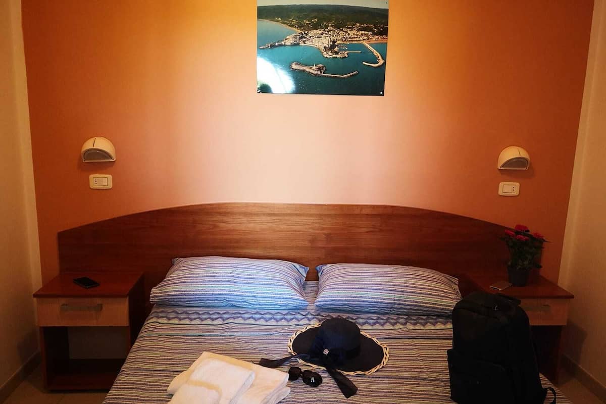 Une chambre d’hôtel avec une tête de lit en bois, deux tables de chevet, un lit avec une literie à rayures, une petite table de nuit avec un téléphone, un chapeau, un sac et un tableau accroché au mur représentant une ville côtière.