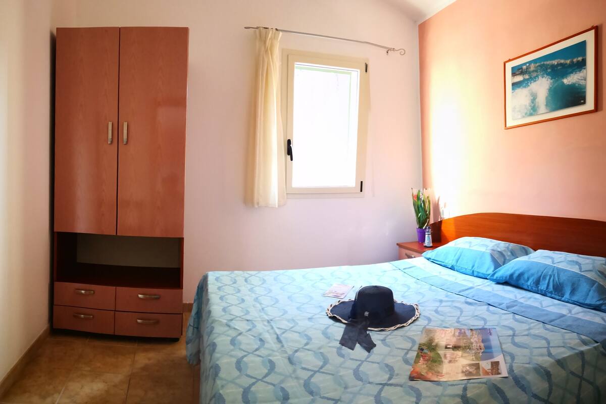 Une chambre à coucher avec un lit, une armoire, une fenêtre avec des rideaux, une table de chevet, une plante et un tableau encadré accroché au mur.