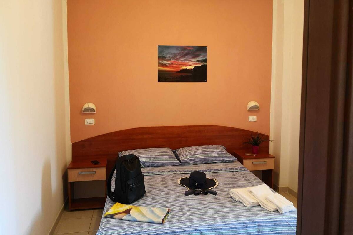 Une chambre d’hôtel avec un lit, une table de chevet, une lampe fixée au mur, un tableau, une valise, un chapeau, des lunettes de soleil et une serviette pliée.