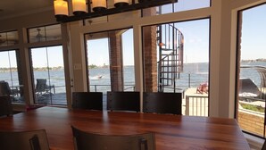 Un comedor con una mesa y sillas de madera, ventanas amplias con vista al agua y un puente, y una escalera que da al exterior.