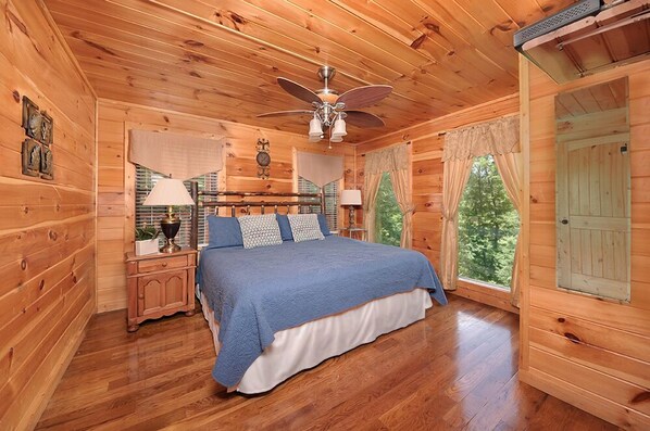 A wooden cabin bedroom with a bed, ceiling fan, and large windows. - 💦Skinny Dippin🌲Private Indoor Pool, Hot Tub, Theater & Game Room (Sevierville)
