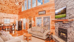 A spacious living room with a fireplace, a large TV mounted on a stone wall, and a cozy seating area with a sofa and chairs. - 💦Skinny Dippin🌲Private Indoor Pool, Hot Tub, Theater & Game Room (Sevierville)