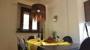 Un coin repas avec une table en bois, des chaises, une nappe jaune, un panier de fruits, une bouteille de vin et une horloge murale.