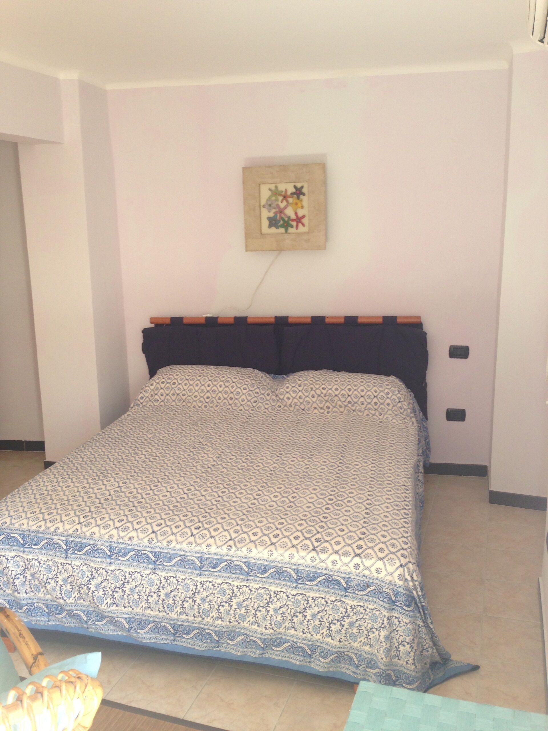 A bedroom with a patterned bedspread, a headboard, a wall-mounted artwork, and a bedside table with a lamp.