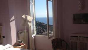 A bedroom with a bed, a chair, a desk, and a window with a view of the sea.