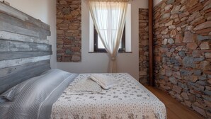 A bedroom with a stone wall, a wooden headboard, a bed with a patterned bedspread, and a window with sheer curtains. - Farmhouse with swimming pool 2 km away from the sea (Piombino (LI))