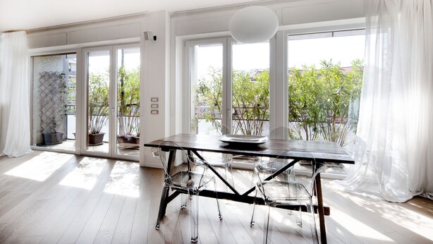 Un comedor moderno con una mesa de madera, sillas transparentes y ventanas amplias que ofrecen vistas a zonas verdes y edificios.