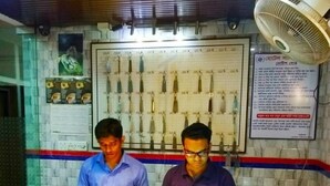 Two men at a counter with documents, a fan above, and a wall with keys and a sign. - Hotel Shalimar Int'l (Dhaka)