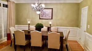 Uma sala de jantar com uma mesa posta para seis pessoas, rodeada por cadeiras, um candelabro e um quadro na parede.