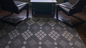 Quarto com uma grande janela com vista para um corpo d'água, duas cadeiras, uma mesinha e um tapete estampado.