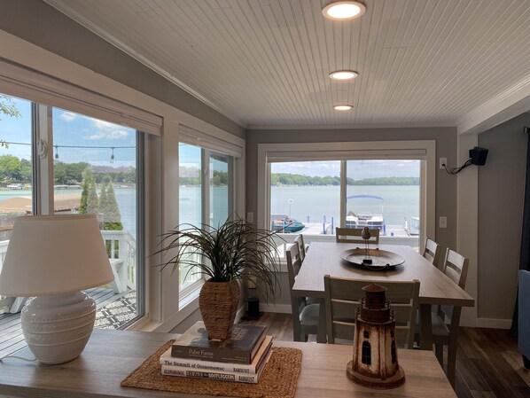 A dining area with a view of a lake, a table with chairs, a lamp, a potted plant, and a rustic lantern. - Waterfront Lake Cottage - Upper Nemahbin Lake (Oconomowoc)