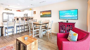 A modern kitchen with white cabinets, a dining area with a wooden table and chairs, a red cabinet with glass doors, and a large window with a view of the ocean. - Pet Friendly Top Floor Gulf View • Free Beach Service • 2 Pools (Miramar Beach)