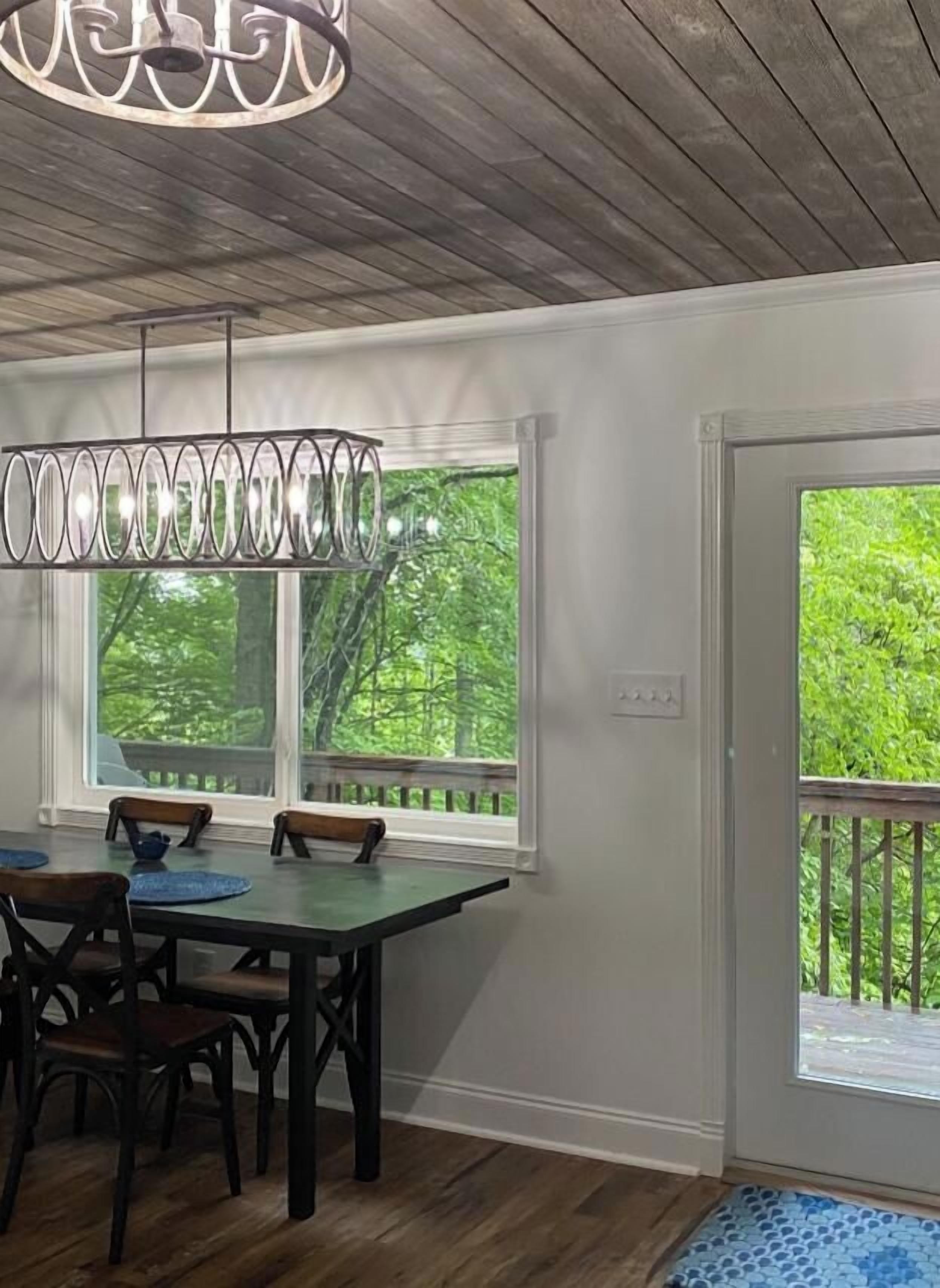 A dining area with a table and chairs, a hanging light fixture, and a view of greenery through the windows.