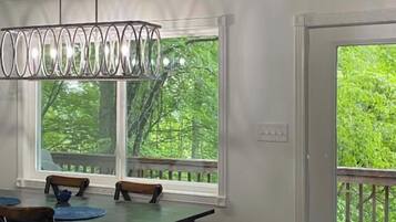 A dining area with a table and chairs, a hanging light fixture, and a view of greenery through the windows.