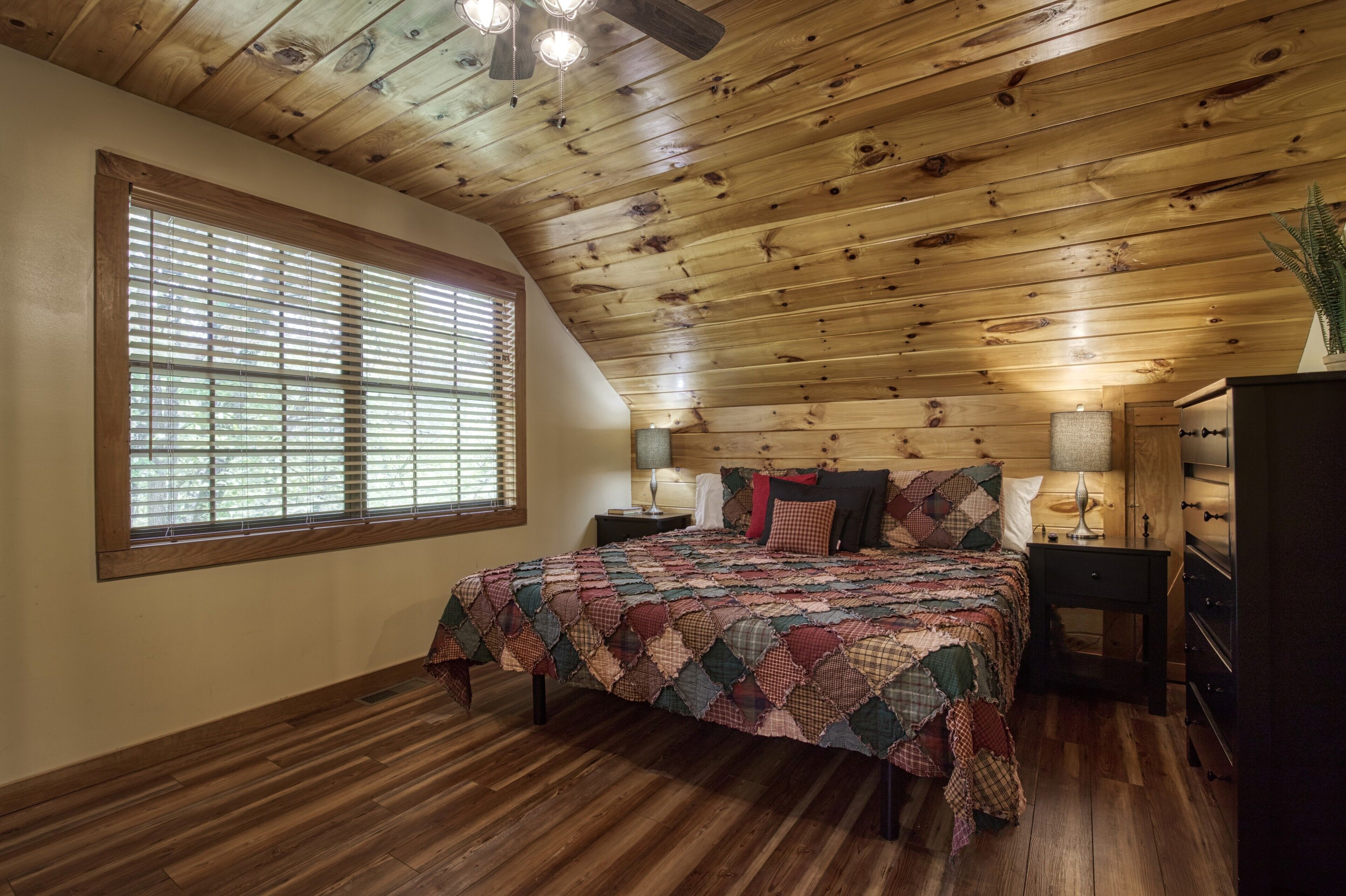 A bedroom with a wooden ceiling, a large window with blinds, a bed with a quilt, bedside tables with lamps, and a dresser.