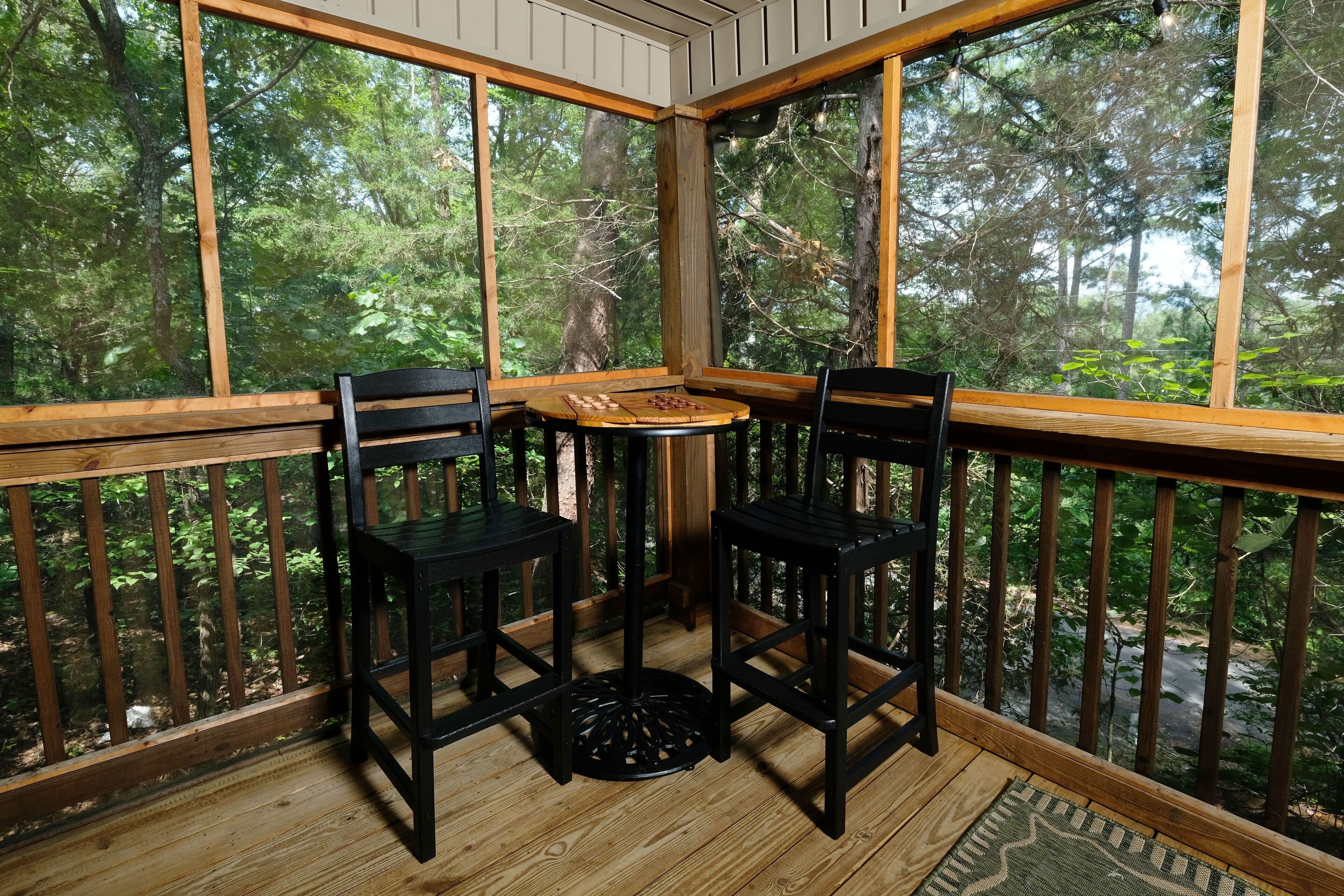 Een houten terras met een tafel en twee stoelen, omgeven door grote ramen met uitzicht op een bebost gebied.