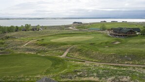Un campo de golf con un club de campo, un lago al fondo y un paisaje montañoso.