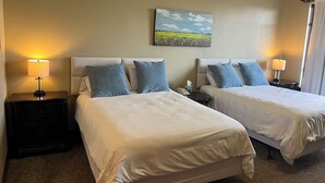 Habitación de hotel con dos camas, cada una con cubrecama blanco y almohadas azules. Hay un cuadro en la pared sobre las camas, una mesita de noche con lámpara, y una pequeña mesa con teléfono y pañuelos.