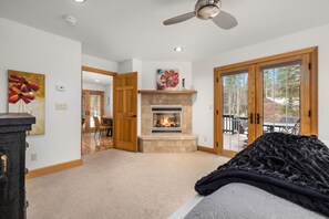 A bedroom with a fireplace, a large bed, a ceiling fan, and a view of the outdoors.