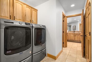 A laundry room with Maytag washers and dryers, wooden cabinets, and a view into a kitchen area. - Blue Star Creekside Lodge  (Columbia Falls)