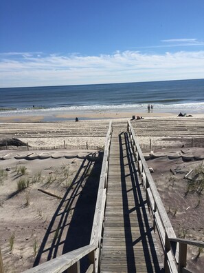 A wooden boardwalk leading to a beach with a clear view of the ocean and a sandy area with some vegetation.