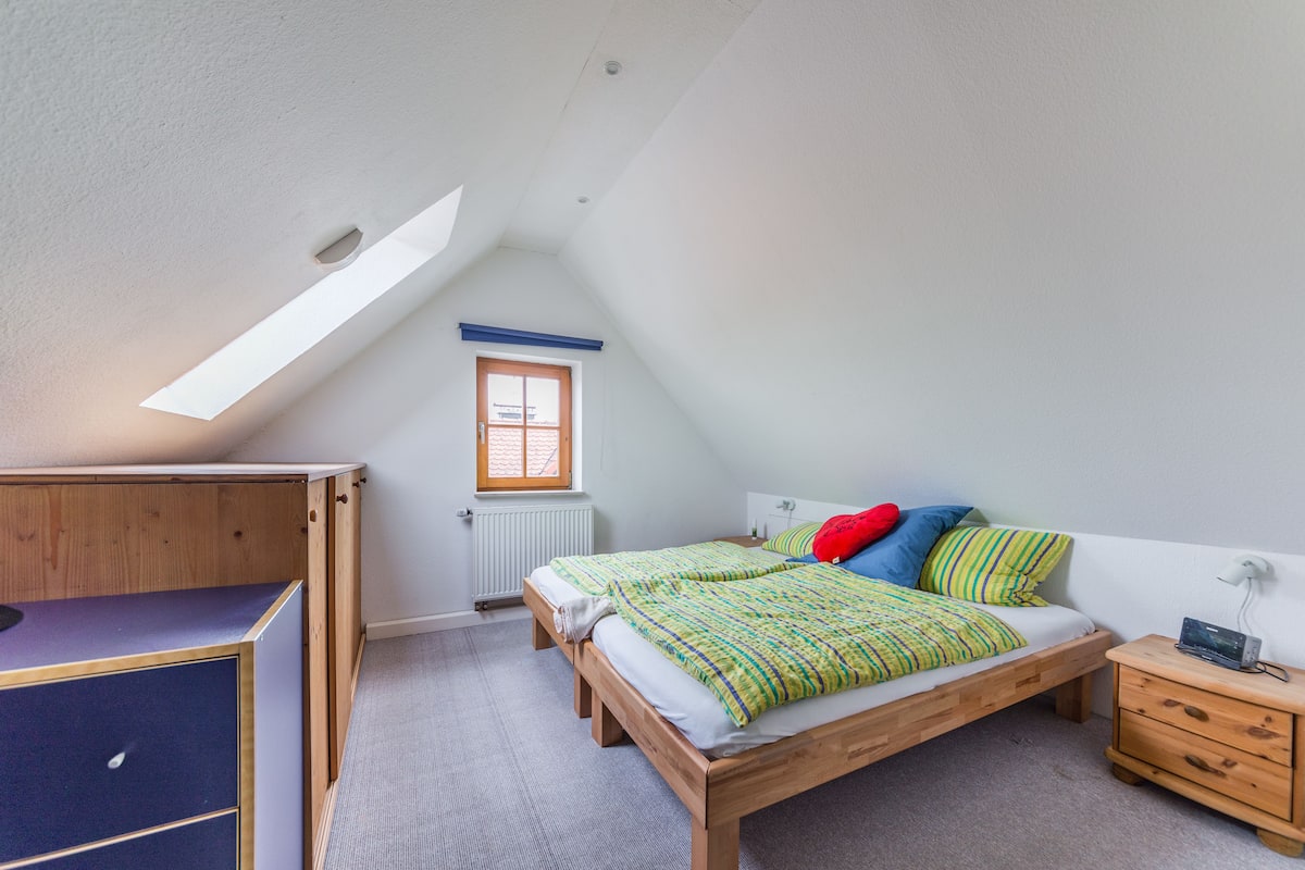 A bedroom with a sloped ceiling, a wooden bed, a nightstand, and a window with a view of the outside.