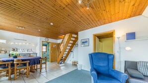 Un vaste espace de vie doté d’un plafond en bois, d’une table à manger avec des chaises, d’un fauteuil bleu, d’un canapé et d’un escalier menant à une autre pièce.