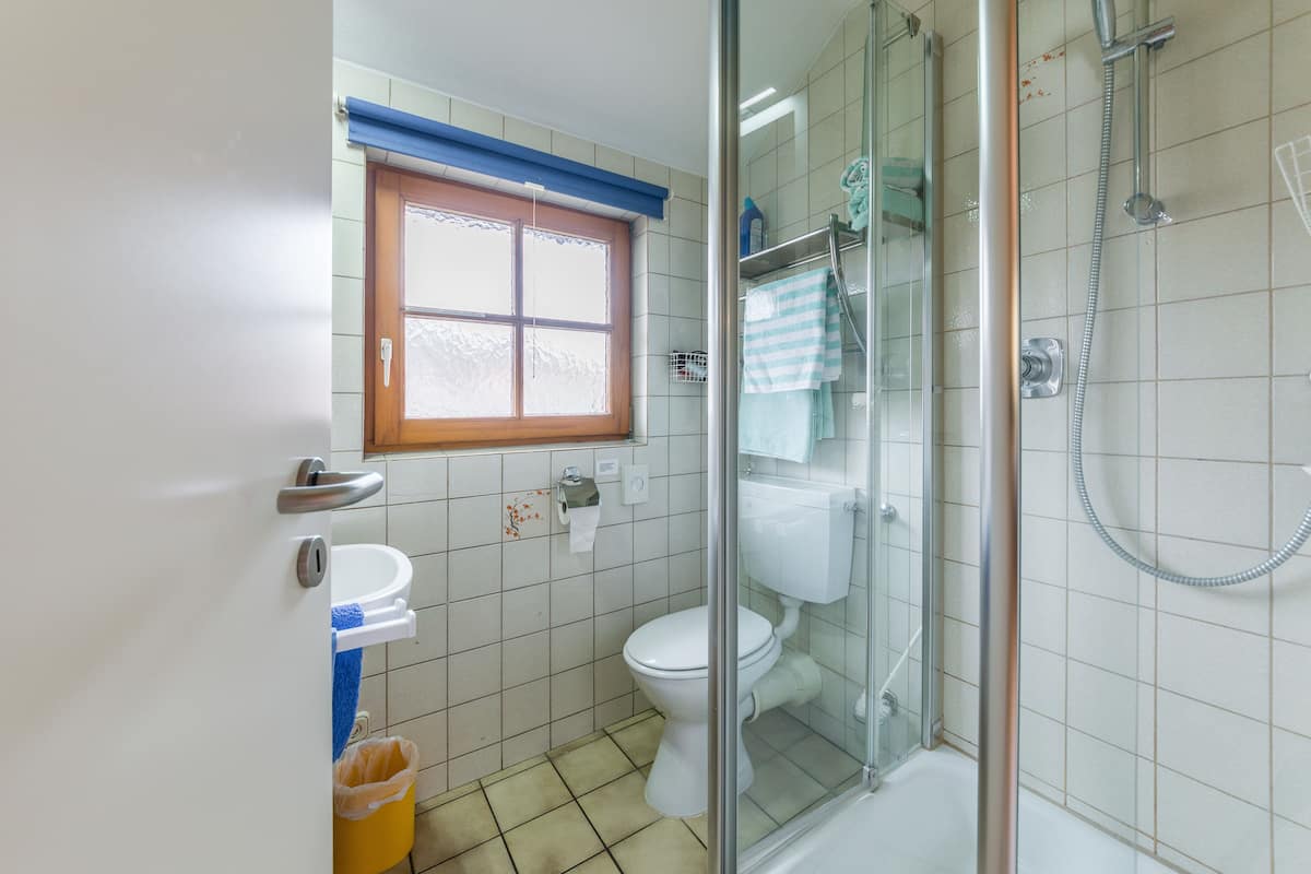 A bathroom with a toilet, a glass-enclosed shower, a window with blue blinds, and a yellow trash bin.