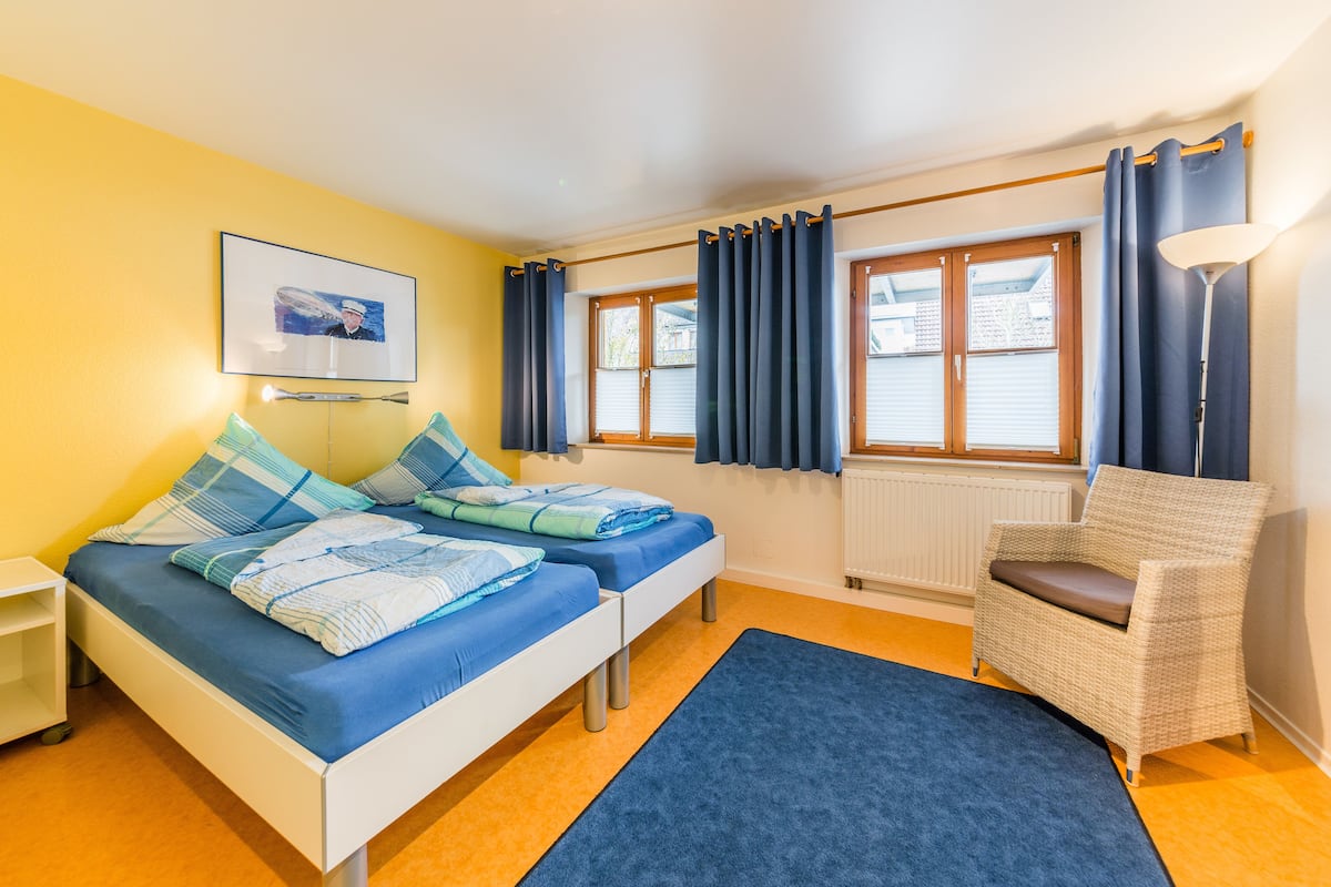 A room with two beds, a chair, a window with curtains, a radiator, and a picture on the wall.