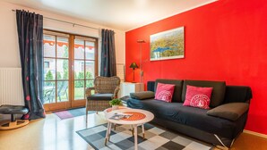 Un salon avec un canapé noir, une table basse blanche, un fauteuil en osier et une vue sur l’extérieur à travers une porte vitrée.