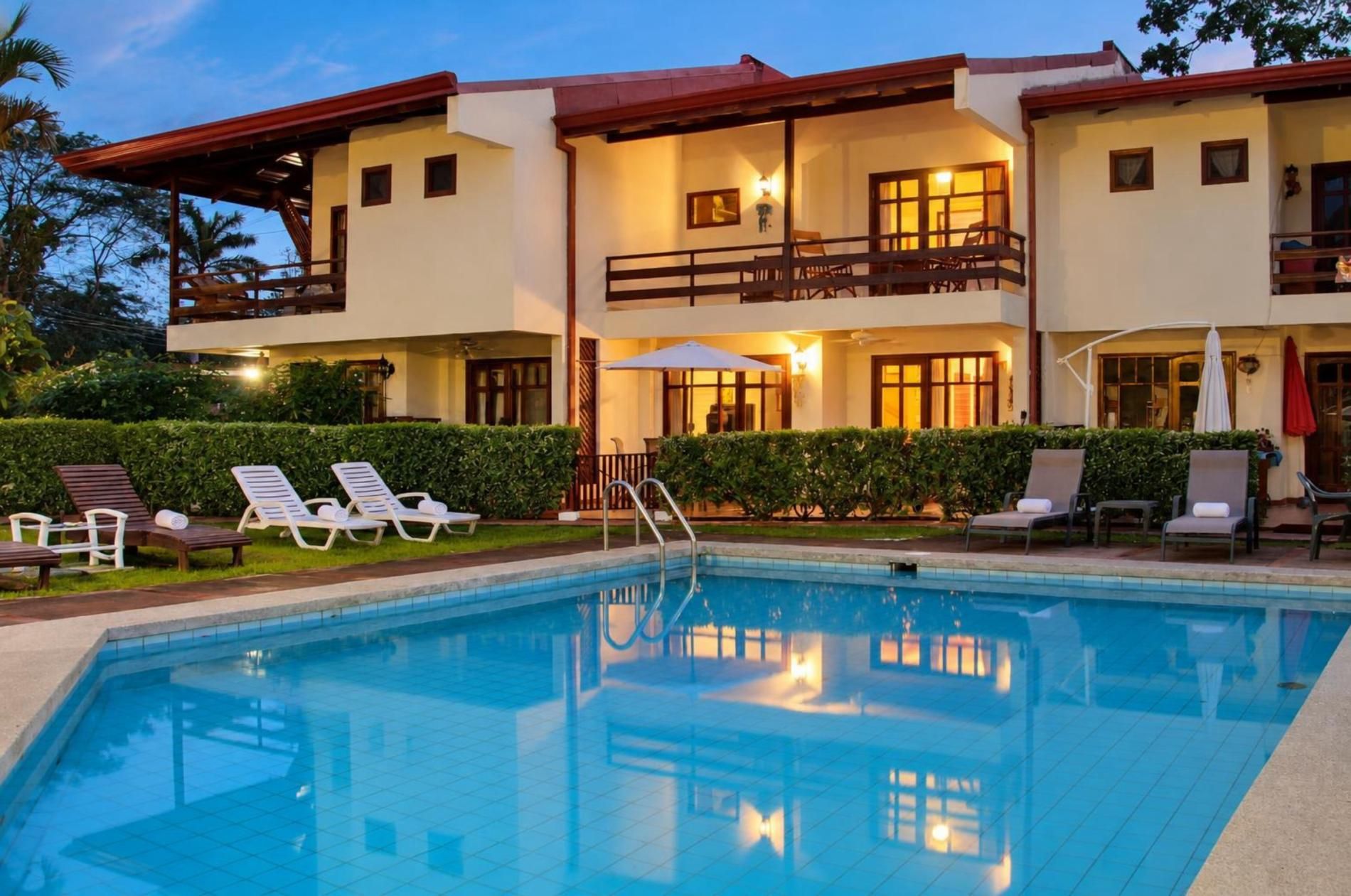 Un edificio de dos pisos con piscina, mobiliario exterior y un balcón bien iluminado.