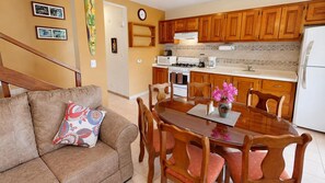A living room with a brown sofa, a dining table with six chairs, wooden cabinets, a kitchen with a stove, microwave, and refrigerator.