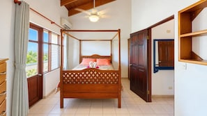 A four-poster bed with a wooden frame and a decorative headboard, a ceiling fan, and a view through the window.