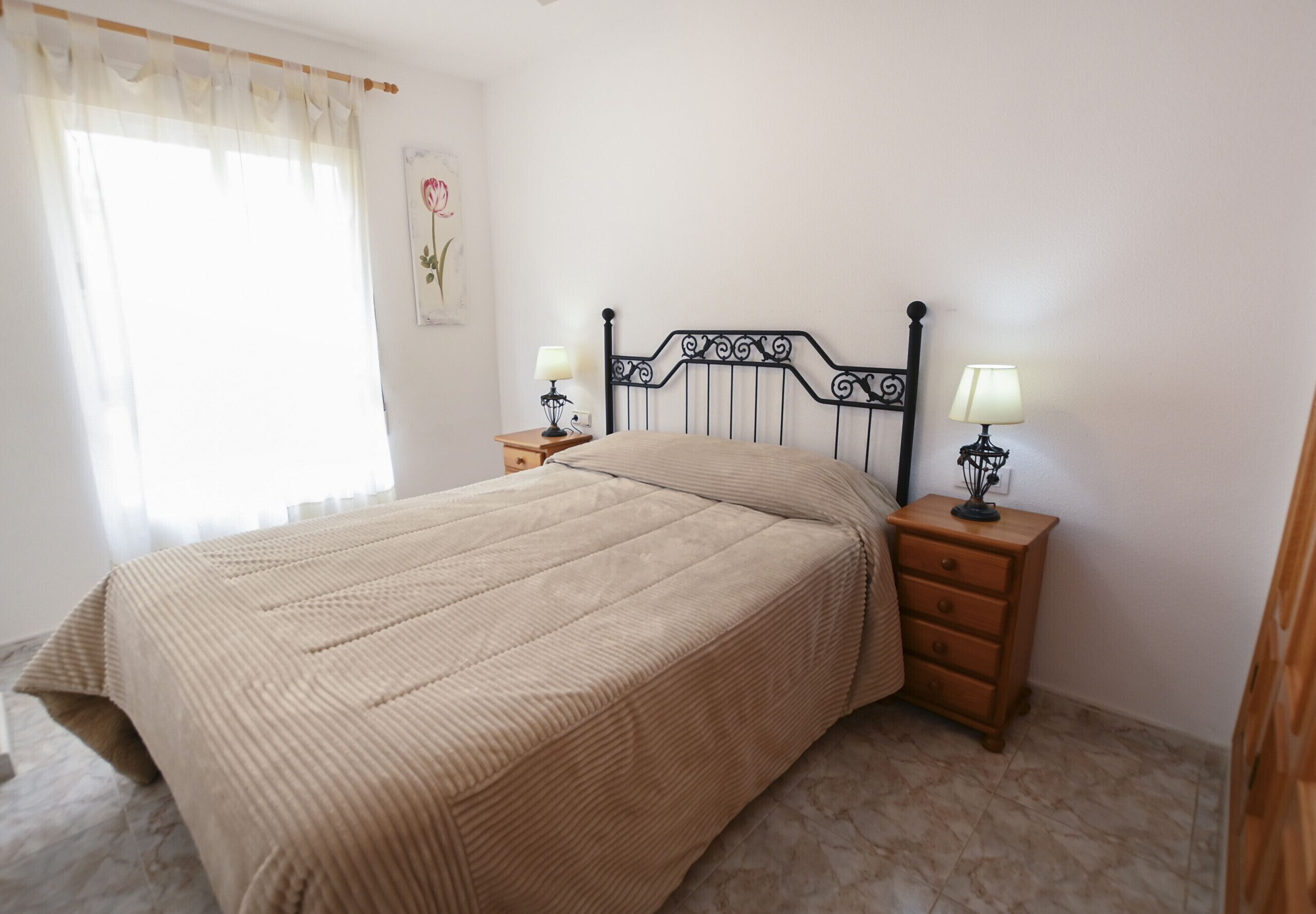 A bedroom with a bed, two bedside tables with lamps, a wooden nightstand, a wall-mounted lamp, and a framed picture.