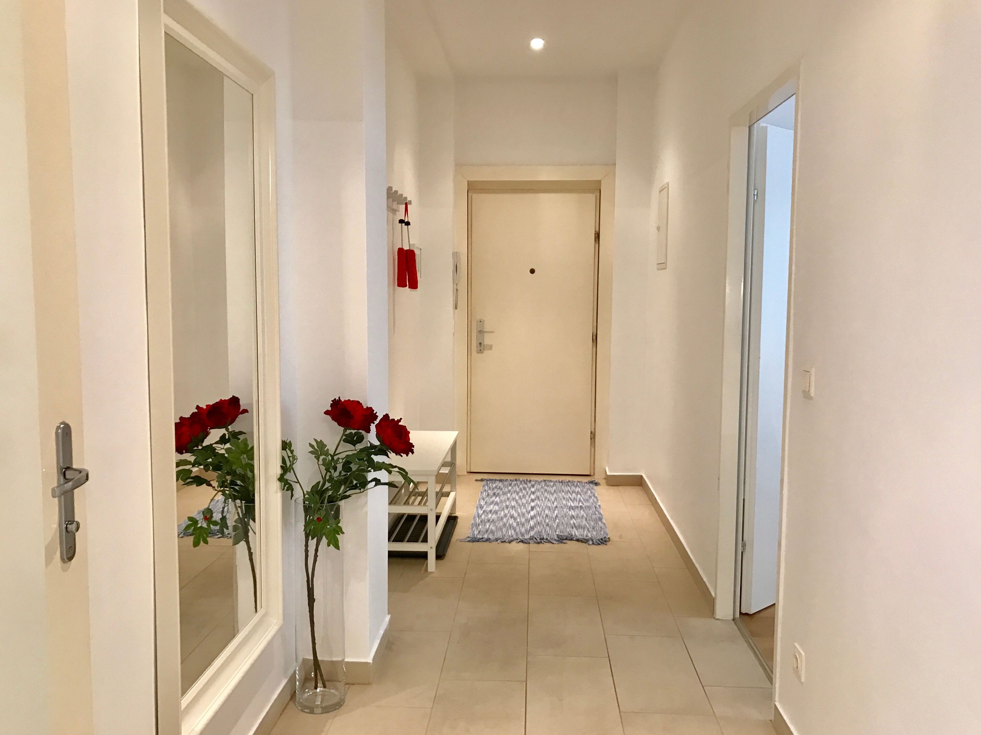 Un couloir avec une porte blanche au fond, une petite table surmontée d’un tapis bleu et blanc, et une plante en pot ornée de fleurs rouges.