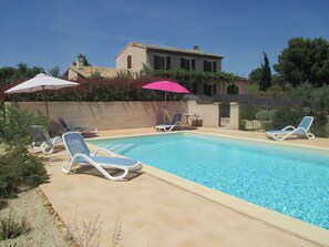 Una piscina amb gandules i ombrel·les, envoltada d'un jardí ben cuidat i un edifici al fons.
