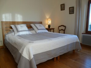 A neatly made bed with a wooden headboard, a bedside table with a lamp, a chair, and a window with curtains.