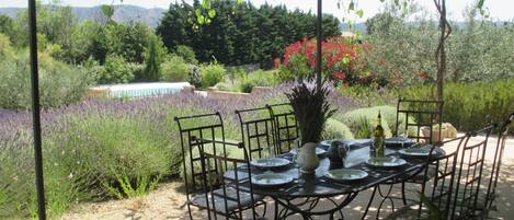 An outdoor dining area with a table set for four, surrounded by chairs, under a pergola with a view of a pool and garden.