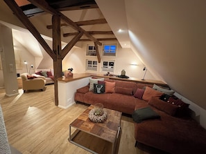 A loft-style living space with a wooden ceiling, a large sofa, a coffee table, and a kitchen area.