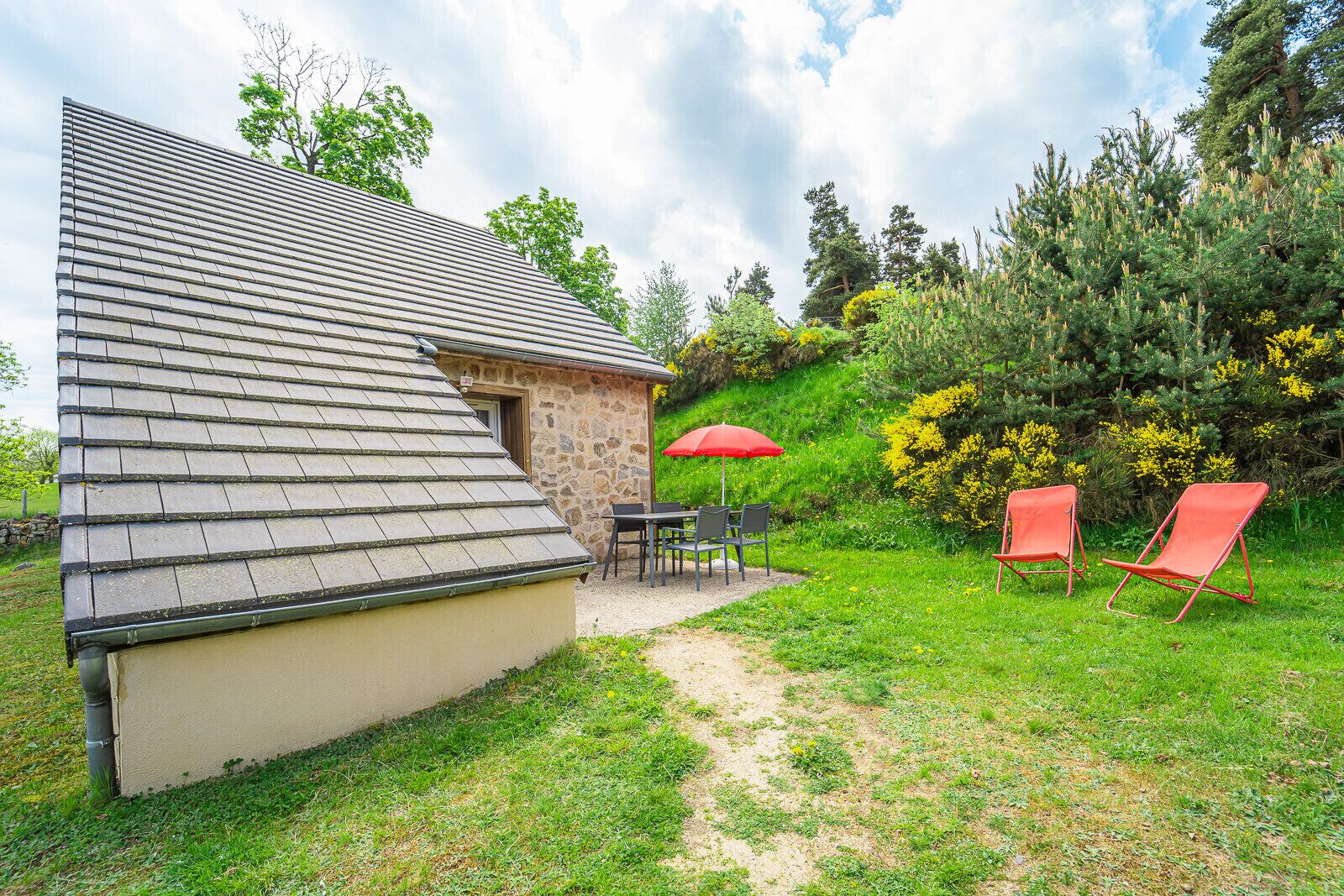 A house with a sloped roof and a stone exterior. There is a patio with a table and chairs, and two red lawn chairs.