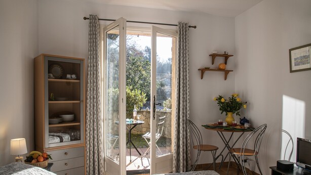 A bedroom with a bed, a wardrobe, a desk with a chair, a small table with a vase of flowers, and a window with curtains.