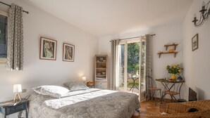 A bedroom with a large bed, a nightstand, a dresser, a chair, a small table with a vase, and a balcony with a table and chairs.