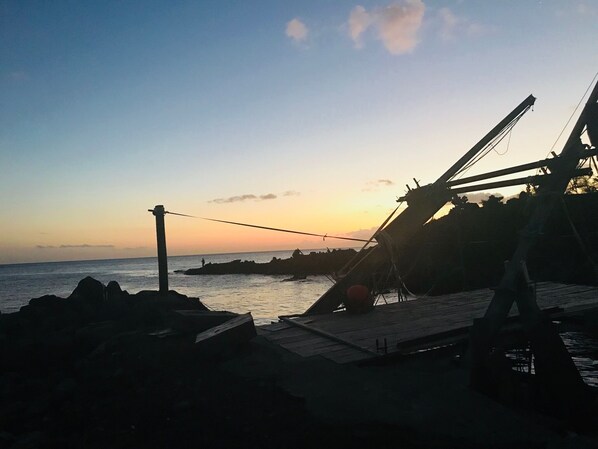 Eine Küstenlandschaft mit einem Steg, der sich ins Wasser erstreckt, einer Person, die auf einem Felsen steht, und einem Sonnenuntergang im Hintergrund.