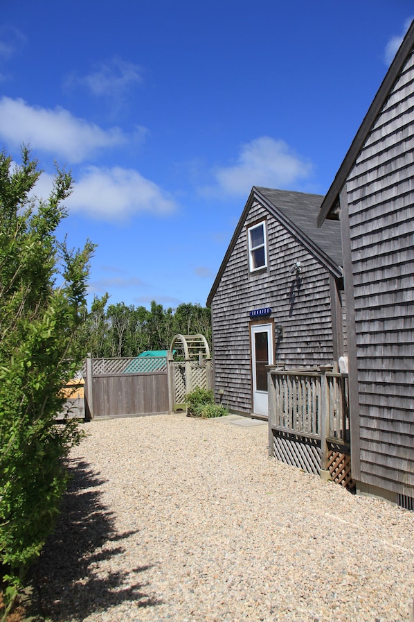 Rustic building with a sloped roof, small window, and wooden door, set amidst a gravel driveway and a tree.