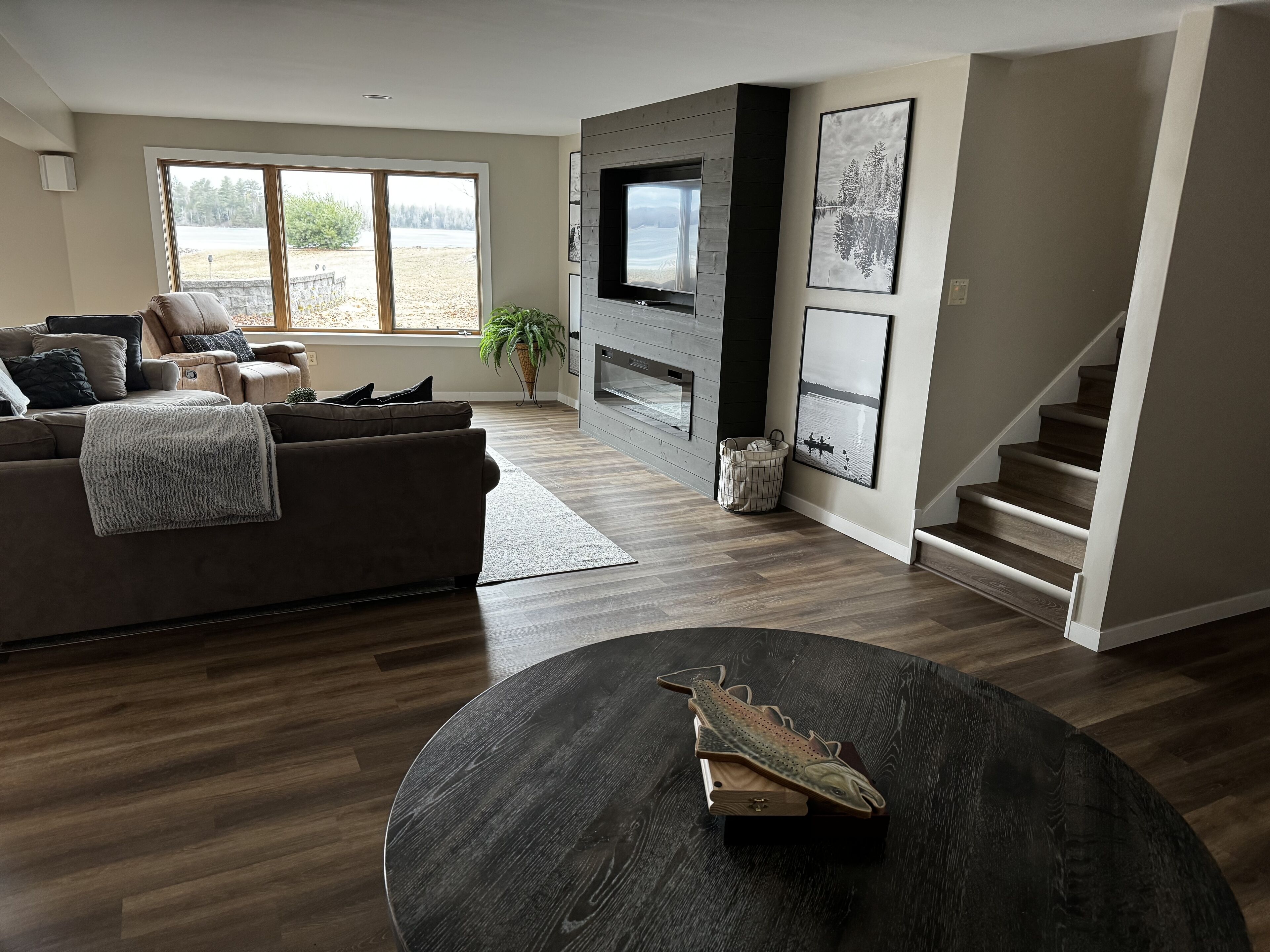 A modern living room with a dark wood floor, a brown leather sofa, a flat-screen TV mounted on a wooden wall, and a staircase leading to another level.